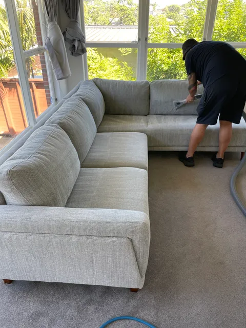 A cleaner using a hand extraction tool on a light fabric corner sofa